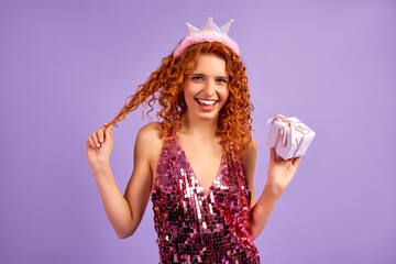 Cute red-haired girl princess with curls in a shiny dress and a crown on her head holding a gift...