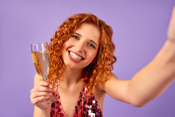 Cute red-haired woman with curls in a shiny dress and a glass of champagne makes a selfie isolated on a purple background. The concept of a party, celebration, birthday.