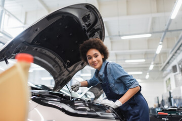 smiling young african american mechanic fixing motor in car with open hood in garage