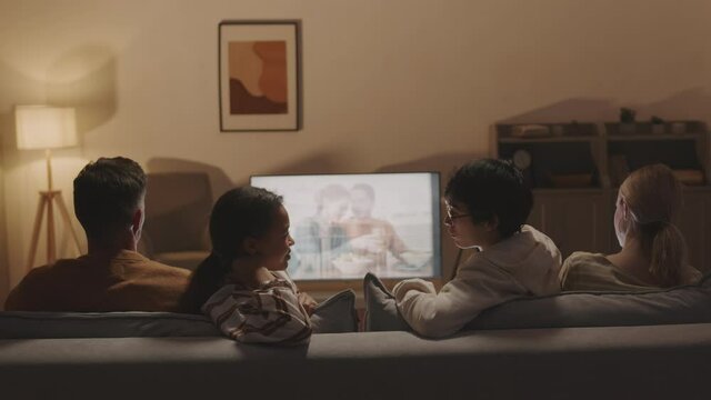 Rear View Of Four Friends Sitting On Sofa In Living Room At Night, Watching Movie On TV, Mixed-Race Woman And Asian Man Talking And Smiling