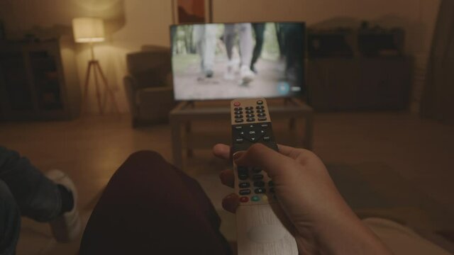 POV of hand of unrecognizable person sitting on couch in living room at night, pointing remote control on TV-set, pushing buttons, turning up volume