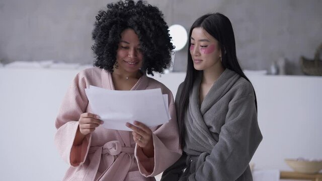 Two Positive Young Women Analyzing Startup Paperwork In Home Office. Portrait Of Beautiful African American And Asian Roommates Planning Business Idea In Bathroom. Slow Motion