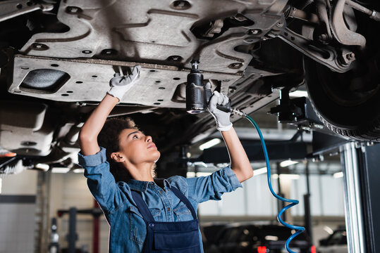 young african american mechanic in overalls repairing bottom of car with electric screwdriver in garage