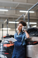Fototapeta premium tired young african american mechanic standing with digital tablet and holding hand near head in garage