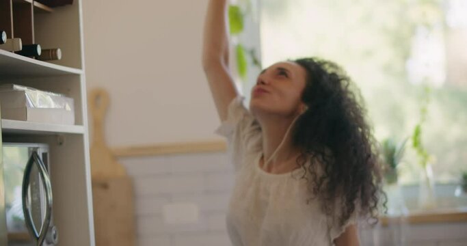 A joyful young woman with curly hair dancing alone in the kitchen, carefree and happy. Slow motion, medium shot, handheld. 
