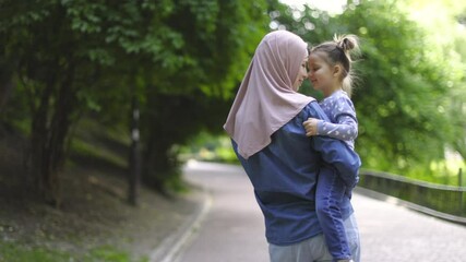 Young pretty happy Muslim mother in hijab, playing with her little adorable daughter in green park and holding her in arms. Slow motion of cheerful family, mom and kid girl, walking in the park - Powered by Adobe