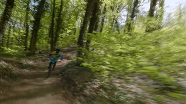 Man rides sportive mtb bicycle along brown ground extreme trail past green trees in bike-park on sunny day. First person fpv drone chase forward view