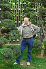 A white-haired fifty-year-old man in jeans and shirt stands in a city park near a tree