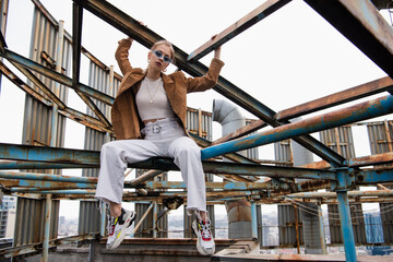 blonde woman in blue sunglasses and suede jacket sitting on rusty construction of rooftop