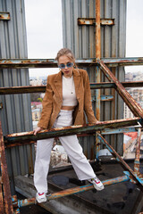 full length of young woman in blue sunglasses and trendy outfit standing on weathered construction of rooftop