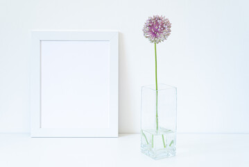Empty white picture frame mockup. Glass vase with allium ampeloprasum flower over white table with white background. Minimal and white interior design. Minimalist and simple decoration