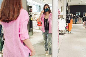 Young woman trying on clothes inside fashion store. Stylish blogger in pink shirt, having fun, wearing face mask. New normal