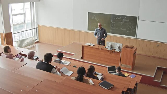 High-angle shot of male mid-adult university professor drawing geometric figures on blackboard teaching students Maths in big auditorium