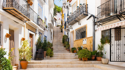 Narrow street with steps, white houses and potted plants in ancient neighborhood El Barrio or Casco Antiguo Santa Cruz in Alicante old town on hillside. Costa Blanca on Mediterranean sea coast, Spain - obrazy, fototapety, plakaty