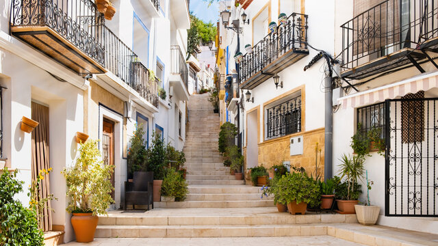 Narrow street with steps, white houses and potted plants in ancient neighborhood El Barrio or Casco Antiguo Santa Cruz in Alicante old town on hillside. Costa Blanca on Mediterranean sea coast, Spain
