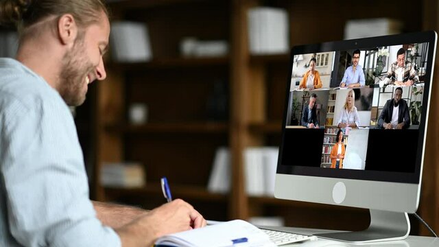 Online education, e-learning. Caucasian smart confident guy sitting at at a work desk, uses computer, listening to an online lecture, taking notes, a teacher and diverse people on the screen