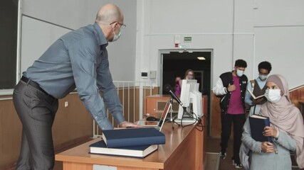 Medium slowmo of bold mid-adult university professor in face mask tracking students attendance on laptop while they walking into auditorium before lesson