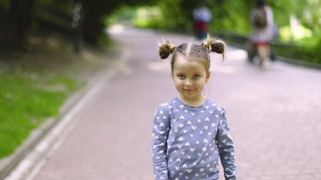 Front view of little pretty child girl, wearing blue shirt, walking and posing on camera in the park outdoors, waving her hand saying hello or goodbye. Slow motion 4K video