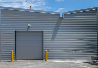 Exterior facade  of large metal industrial building with grey corrugated metal siding, grey garage door, roof with inverted slope, daytime, nobody