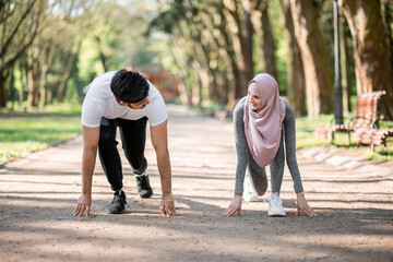 Young muslim couple in sport clothes standing in start position for run at green summer park. Man and woman in hijab preparing for morning workout outdoors.