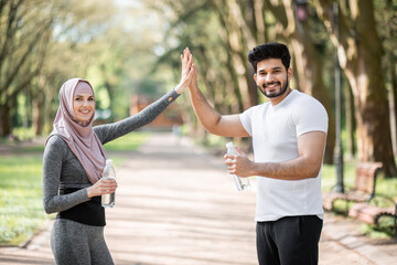 Happy muslim man and woman in hijab giving high five to each other, smiling and looking at camera while resting after training at park. Concept of relationship, sport and lifestyle.