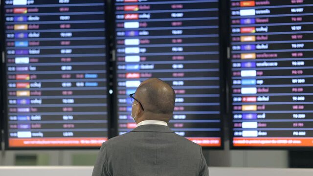 Young businessman with trolley bag in the airport. Wearing mask. traveling during pandemic must be social distancing. He standing at information flight timetable waiting plane at airport terminal.