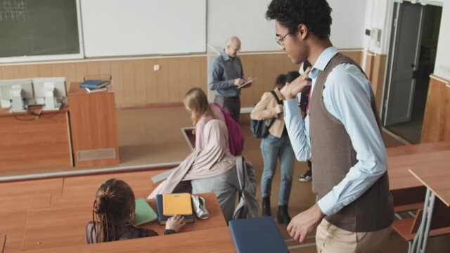 High-angle slowmo shot of group of young multi-ethnic students taking their seats by long wooden desks in bid auditorium greeting each other before beginning of lesson