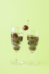 Two glasses of champagne filled with a cherry drink on a green background. One cherry between two glasses as a connection.