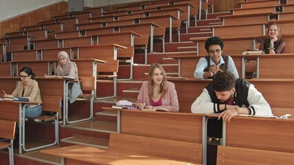 Medium slowmo of young bearded male student in sports bomber jacket leaning on desk sleeping at big auditorium or lecture hall while classmates trying to wake him - Powered by Adobe