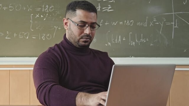 Tilt-up medium slowmo of mid-adult Mixed-race professor of Maths typing on laptop while sitting at desk in modern bright classroom with Geometry problem solution written on blackboard in background
