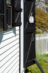 A white and black sign with the words salt life written on it. The sign is hung on a black wooden exterior door of a shed.  The building has a white clapboard exterior wall with a window and shutters.