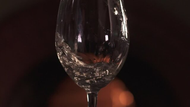 Close-up of a tall transparent glass in which white wine is poured in a dark room against the background of the fireplace.  Bokeh fire in the background. Slow motion 120 fps