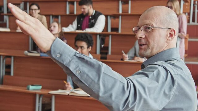 Rear-view waist-up slowmo shot of bold mid-adult professor giving lecture to small group of diverse male and female students pointing at blackboard