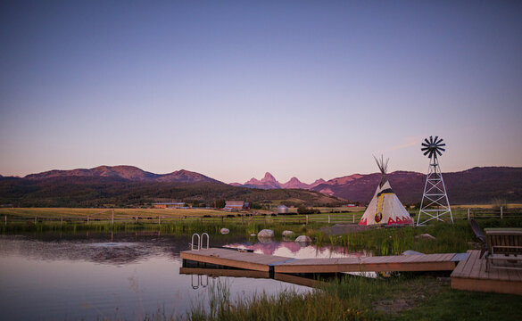 A lake with a dock, a teepee and a windmill with the Tetons in the background on a ranch in Alta, Wyoming, on the west side of the Teton Mountains. 