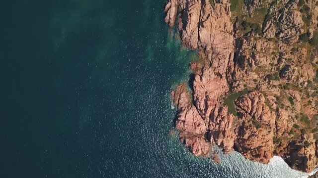 Aerial view of a reddish secluded reef surrounded by a transparent blue sea near Isola Rossa, Sardinia, Italy.
