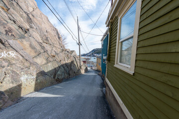 A narrow paved asphalt street with multiple wooden buildings on one side and a large rock mountain on the other. The exterior of the houses is covered in wood clapboard with double hung windows. 