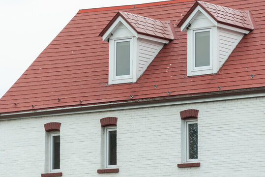 The corner of a red metal shingled roof with two raised dormers and three sunken small single windows in the exterior wall of a white stucco building. The vintage building has a clean design.