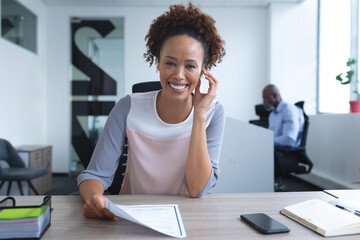 Portrait of mixed race businesswoman sitting at desk smiling with colleague in background