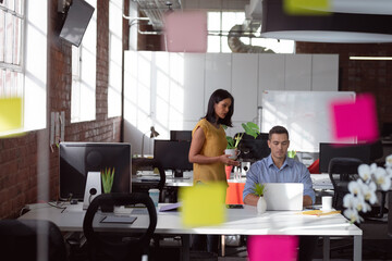 Caucasian male and female colleague in discussion at desk looking at laptop and drinking coffee