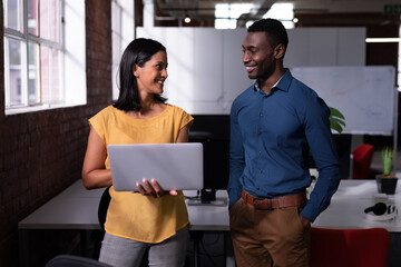 Smiling diverse male and female colleague standing in office looking at laptop together - Powered by Adobe
