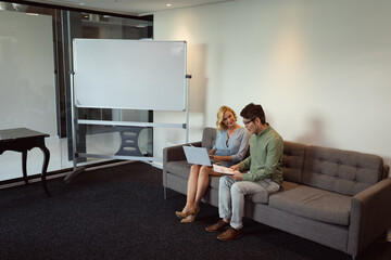 Diverse male and female colleague sitting in office lounge discussing and using tablet and laptop