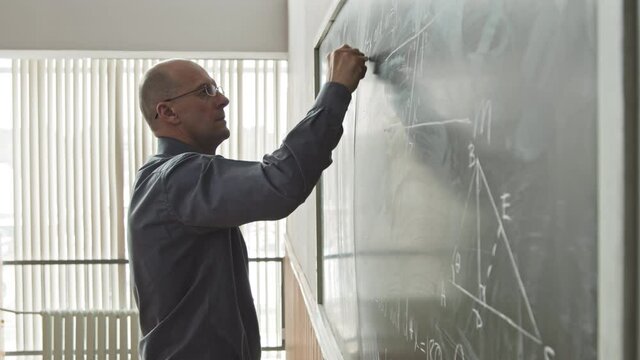 Side-view medium slowmo of bold mid-adult Caucasian university professor of Mathematics in eyeglasses drawing geometric figures on blackboard in auditorium