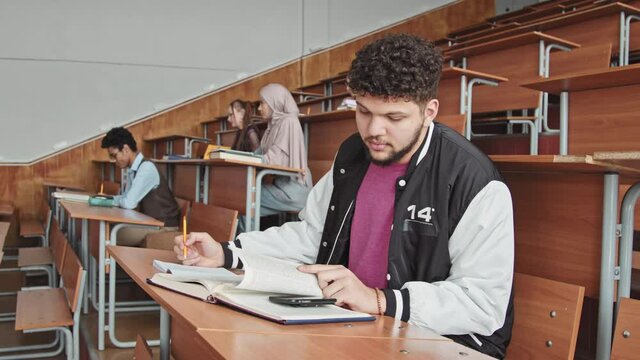 Side-view medium slowmo of young male student sitting by desk in auditorium trying to cheat at exam, looking at smartphone put between pages of student book
