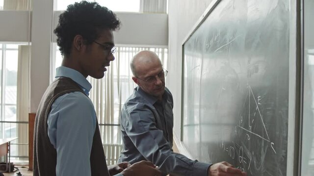 Medium slowmo of contemporary male mid-adult professor explaining his young Mixed-race student how to solve Geometry problem standing together at blackboard in classroom