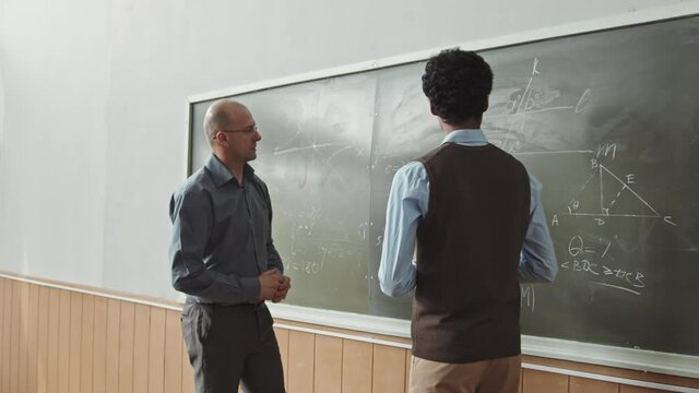 Aerial medium slowmo shot of male Mixed-race student answering at blackboard trying to solve hard Geometry problem with help of mid-adult professor