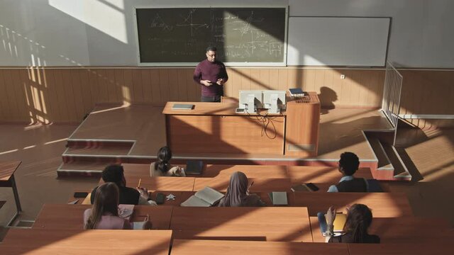 Top-view of high school or university students sitting by long wooden desks in auditorium having Maths lesson, looking at male mid-adult professor standing at blackboard while explaining new topic