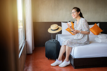 Tourist woman sitting on bed nearly window, looking to beautiful view with her luggage in hotel...