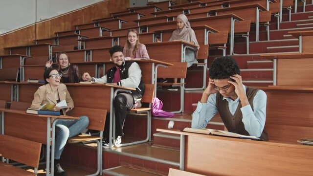Slowmo of group of high school or university male and female students bullying their sad lonely Mixed-race classmate throwing crumpled paper at him while boy sitting separately and reading book