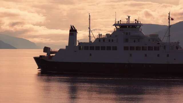 Kanestraum-Halsa ferry crossing. A large ferry carrying cars and passengers across the narrow fjord. The large vessel moving swiftly, leaving low waves in its wake. Pale orange clouds cover the sky