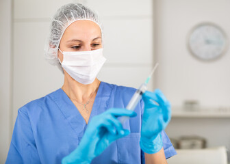 Nurse in a protective mask, working in the hospital, stands with a syringe filled with medicine in the treatment room